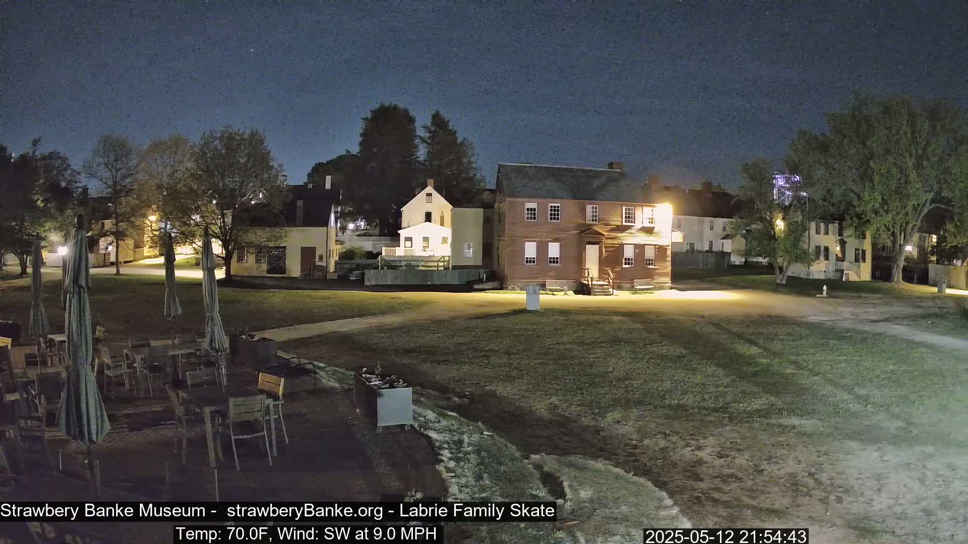 A nighttime view of a grassy area with several buildings, outdoor seating, and umbrellas under a clear, dark sky.