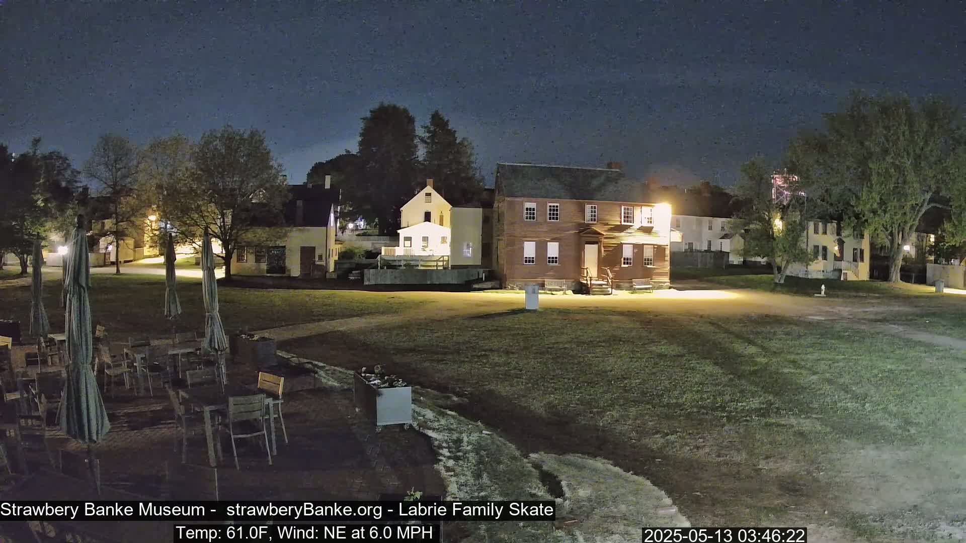 A nighttime view of a grassy area with several buildings, outdoor furniture, and umbrellas under a clear, dark sky, with a gentle northeast wind.