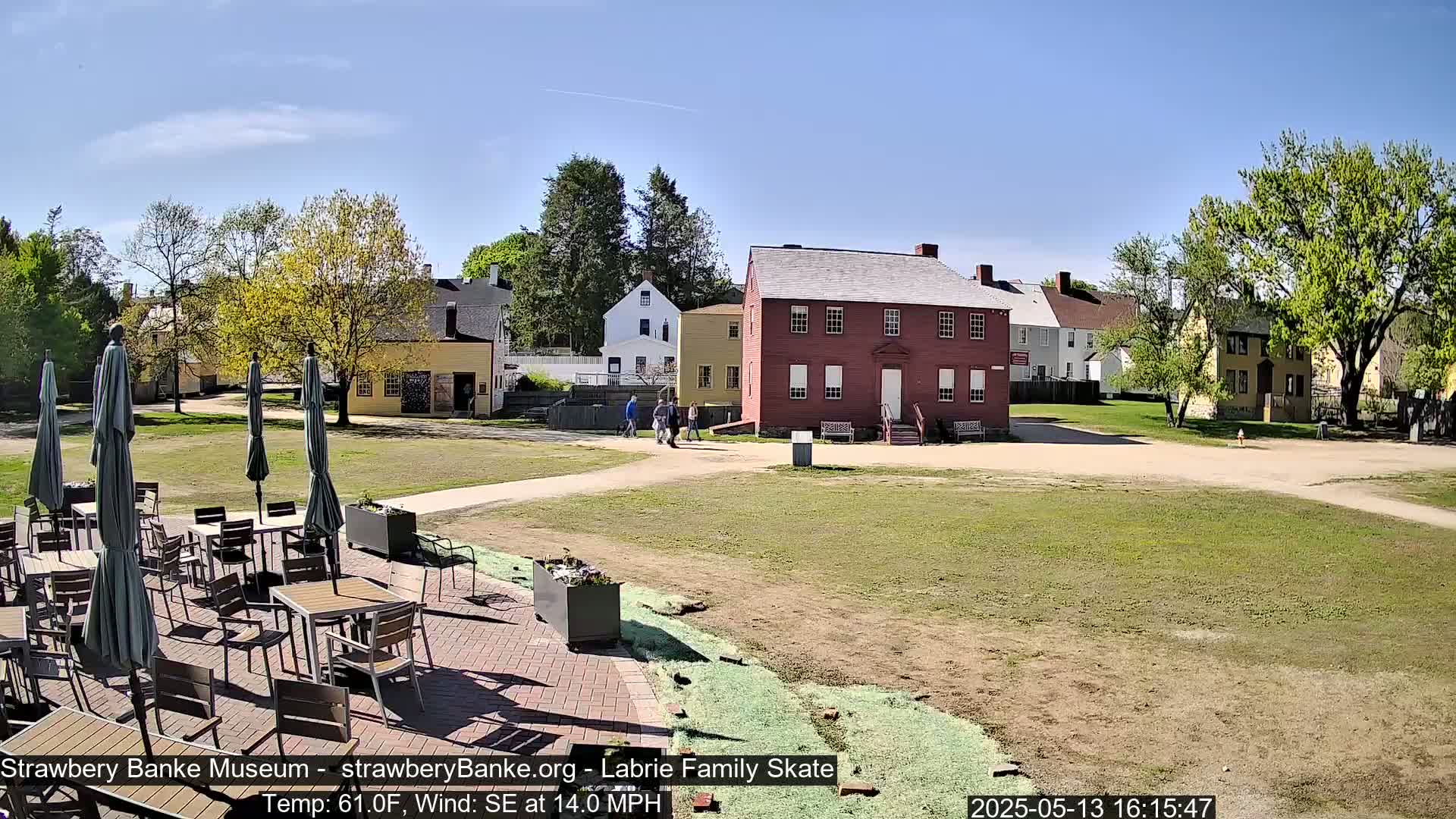 A sunny outdoor patio with tables and umbrellas sits before several historic-looking buildings on a grassy expanse, under a bright blue sky with a light southeast wind.
