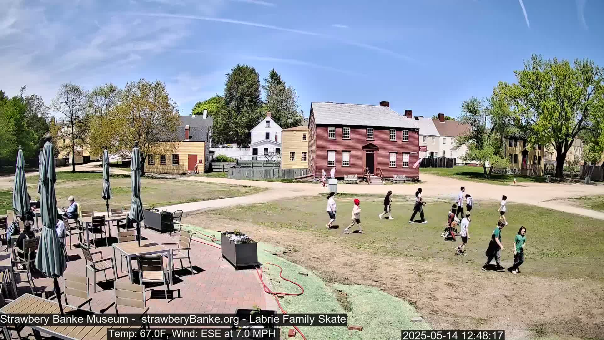 A sunny outdoor scene shows a grassy area with several people walking, children playing, and an outdoor patio seating area with umbrellas and tables near several historic-looking buildings.