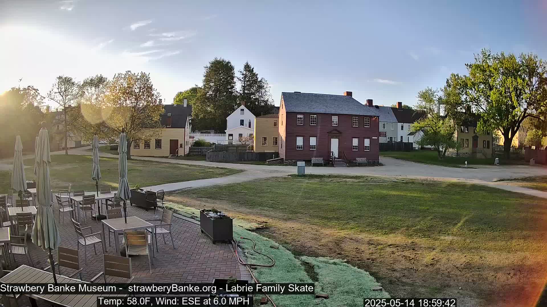 A patio with tables and umbrellas overlooks a grassy area with several historic-looking buildings in the late afternoon sunlight, with a gentle ESE wind.