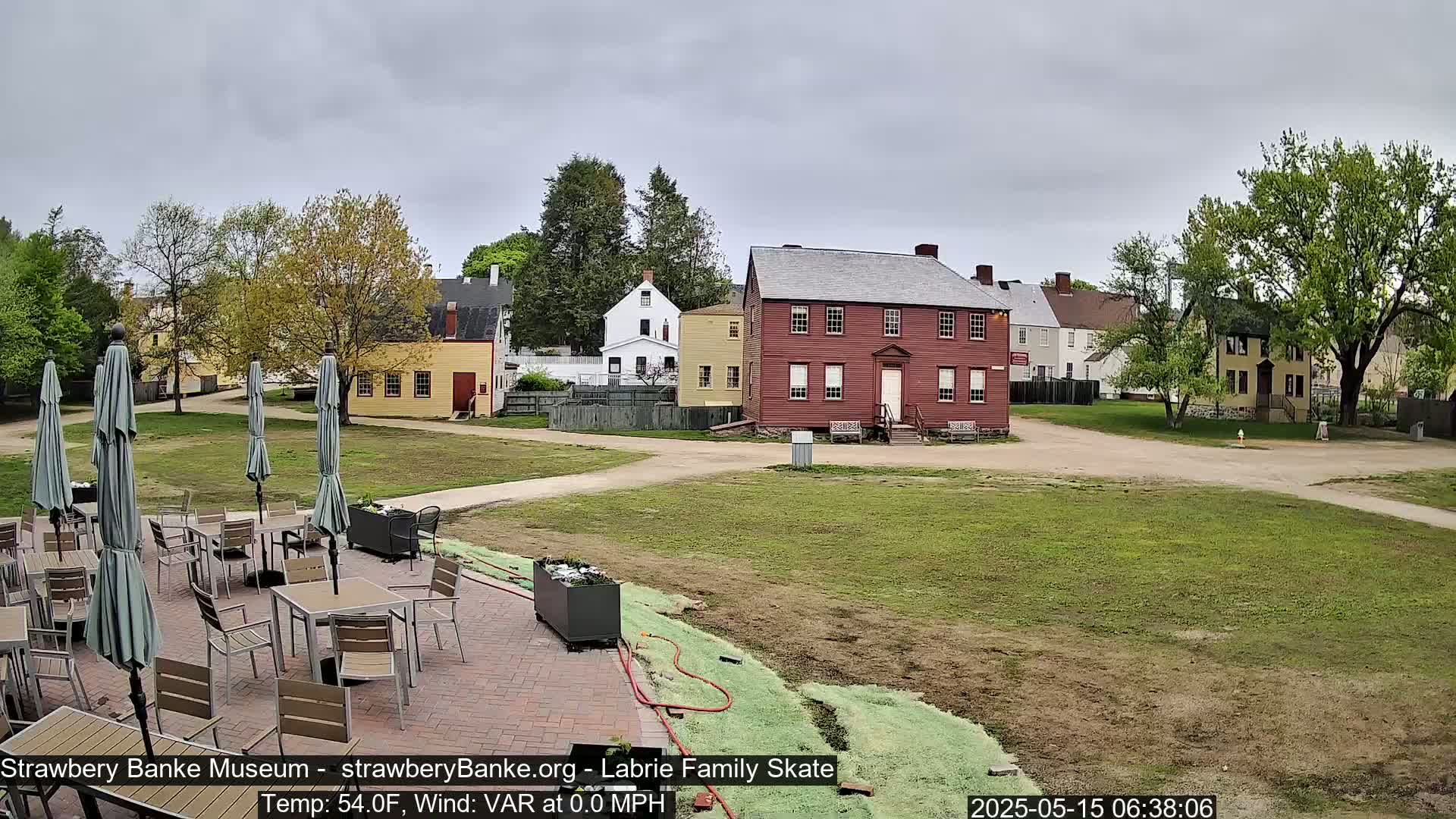 A patio with tables and umbrellas sits in front of several historic-looking buildings on a partly cloudy day.