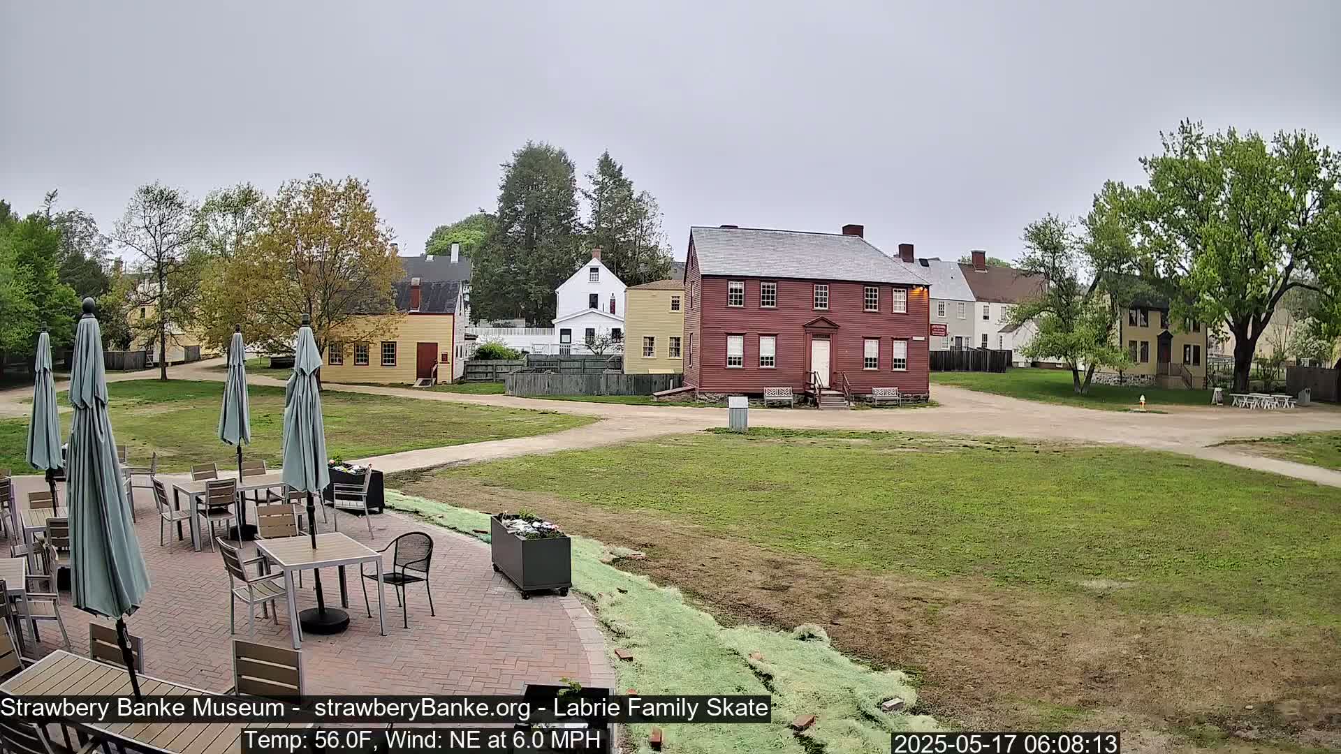 A patio with tables and umbrellas sits in front of a collection of historic buildings on a partly cloudy day with a northeast wind.