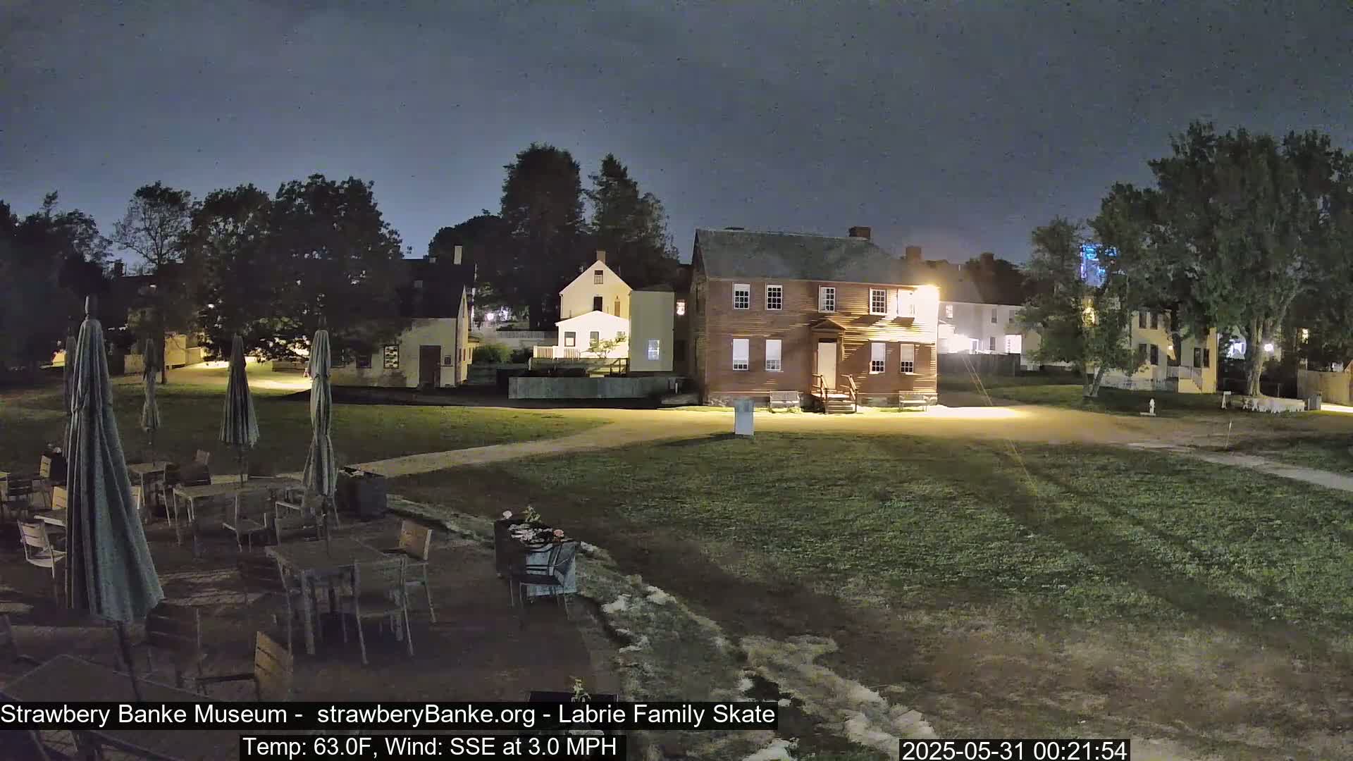 A nighttime view of a grassy area with several tables and umbrellas, situated in front of a collection of historic-looking buildings under a dark sky.