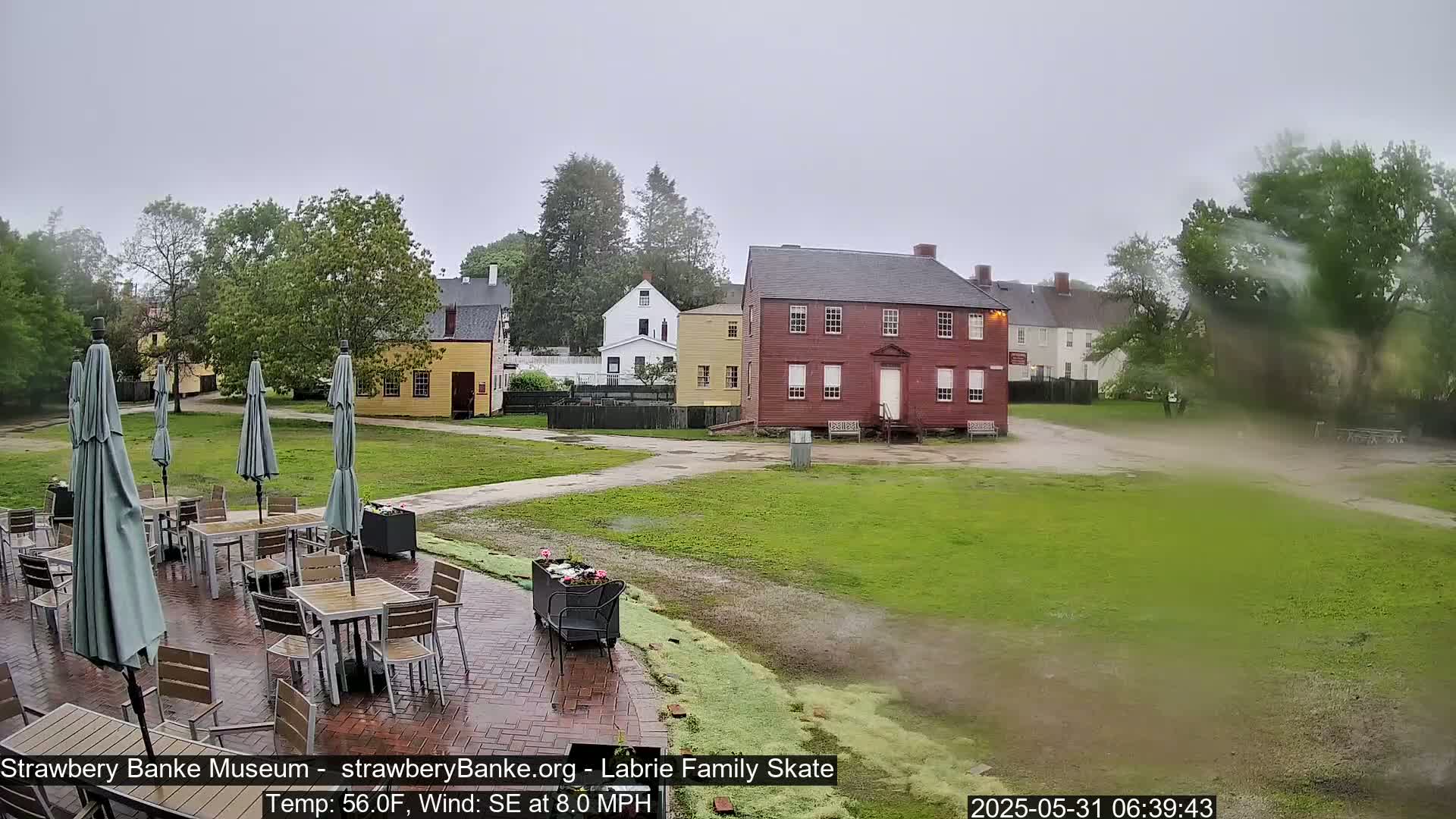 A brick patio with tables and umbrellas sits before several historic buildings on a grassy lawn in rainy, windy weather.