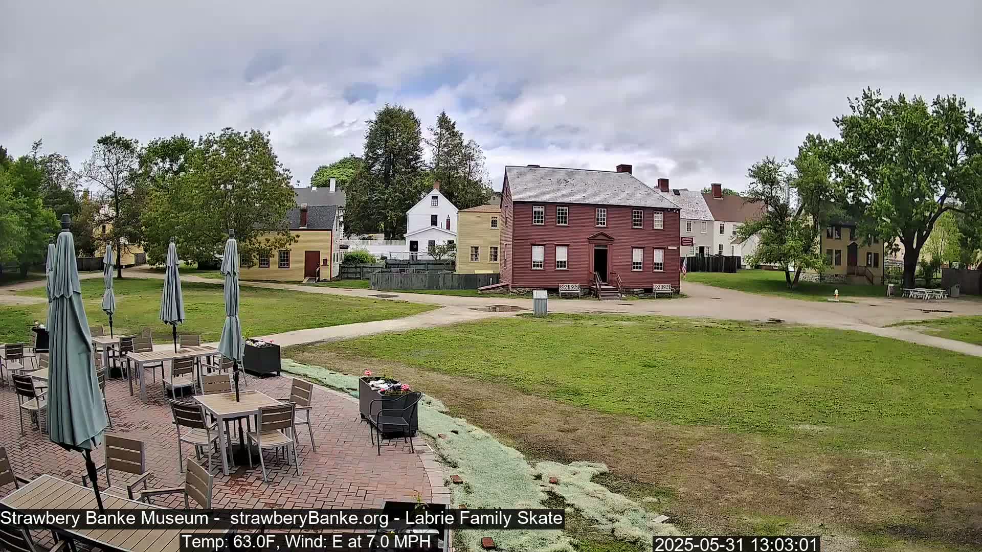 A brick patio with tables and umbrellas sits in front of a grassy area, overlooking several historic-looking buildings under a partly cloudy sky.