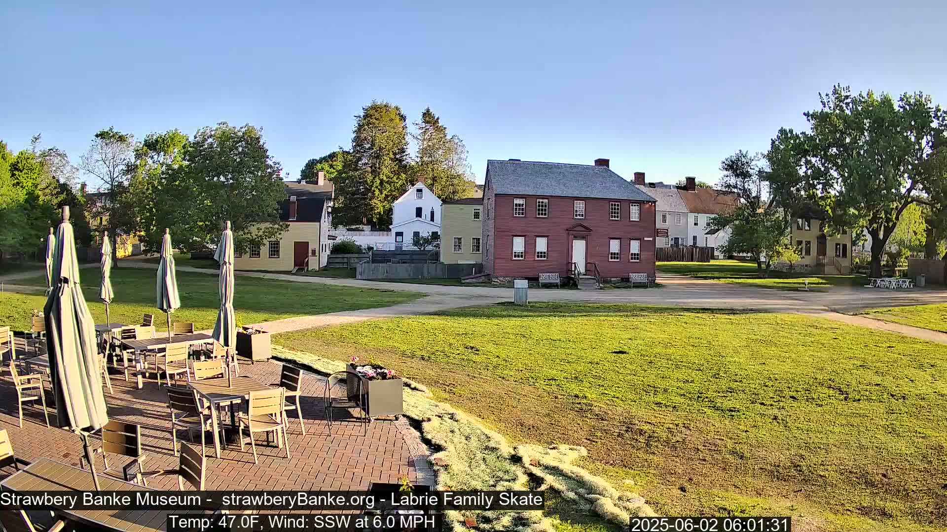 An outdoor patio with tables and umbrellas sits in front of a grassy area and several historic-looking buildings on a clear, sunny morning.