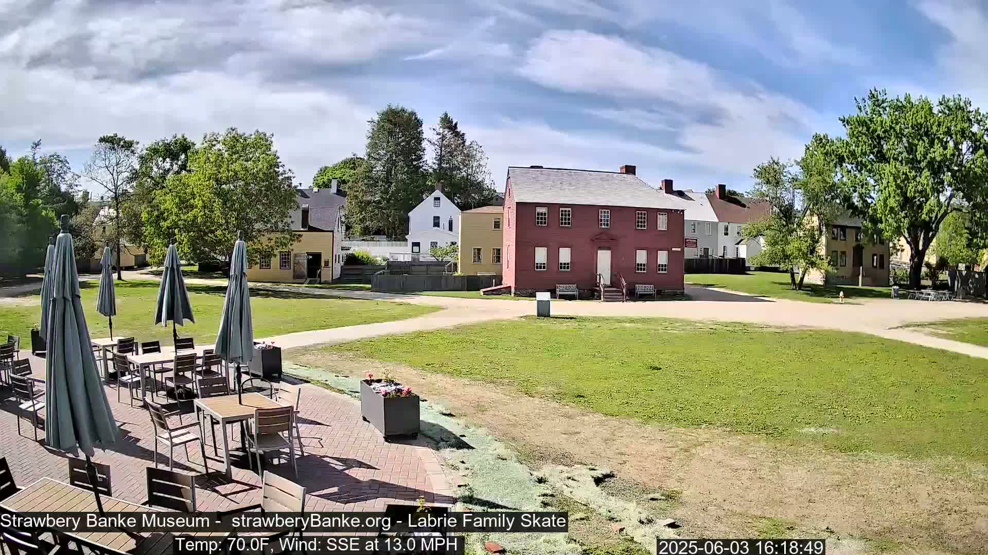 A sunny outdoor scene shows several historic-looking buildings surrounded by grassy areas and a patio with tables and umbrellas.