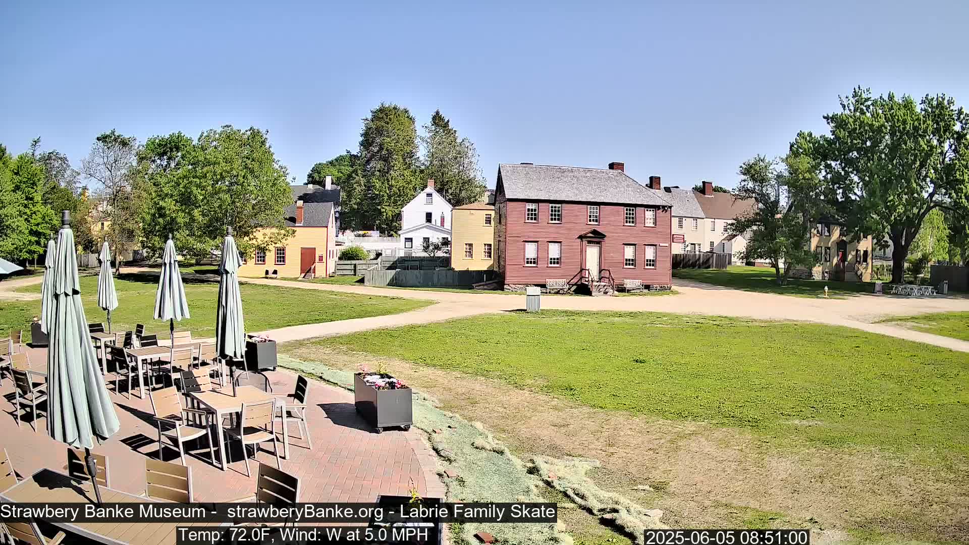 A sunny outdoor scene shows several colonial-era buildings and a patio area with tables and umbrellas.