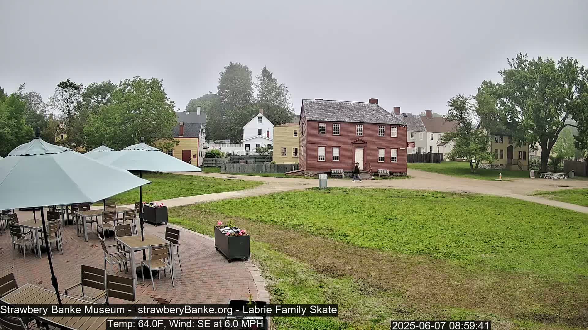 A brick patio with several light-blue umbrellas and tables sits before a grassy area and a collection of historic-looking buildings on an overcast day.