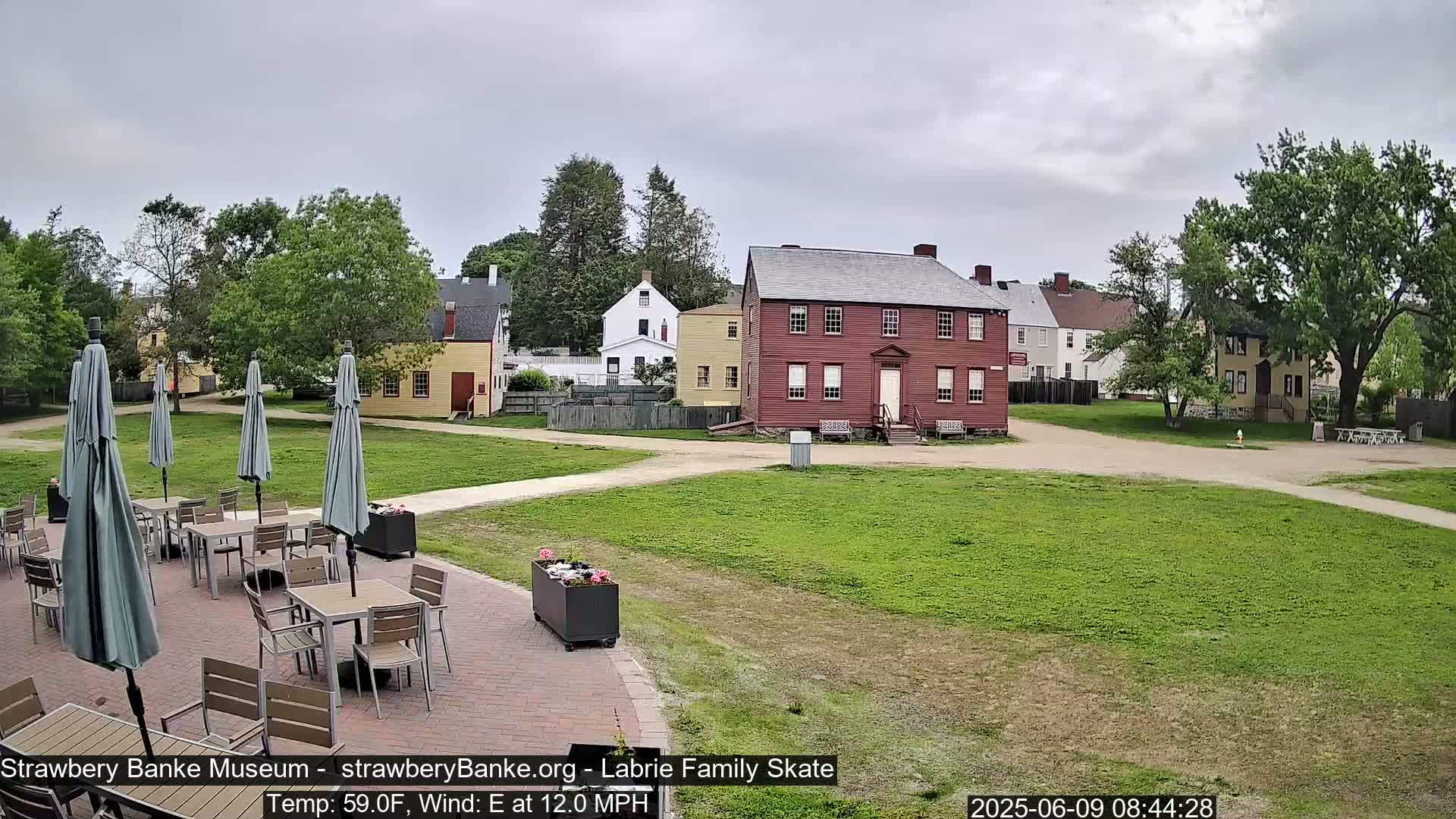 A paved patio with tables and umbrellas sits before a grassy area and several historic-looking buildings under an overcast sky.