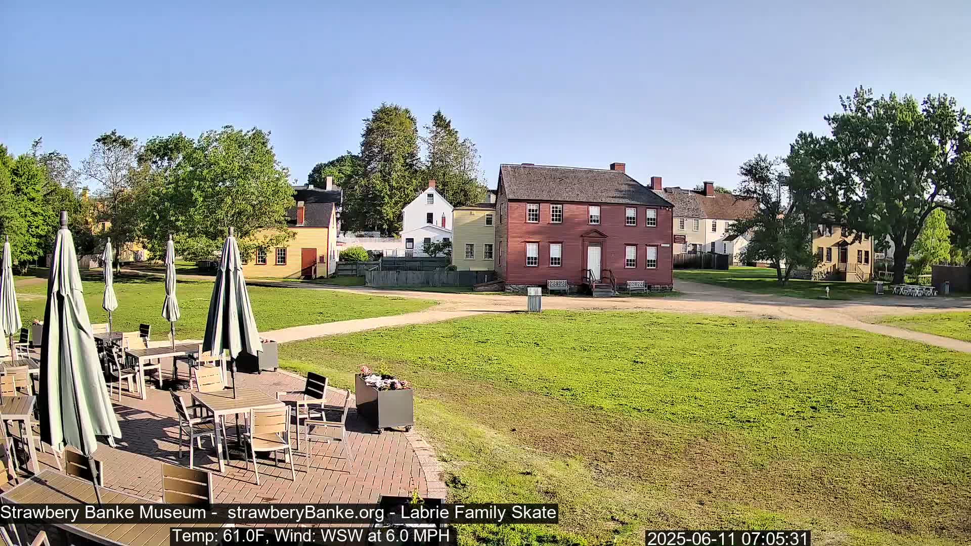 A patio with tables and umbrellas overlooks a grassy area with several historic-looking buildings under a clear, sunny sky.