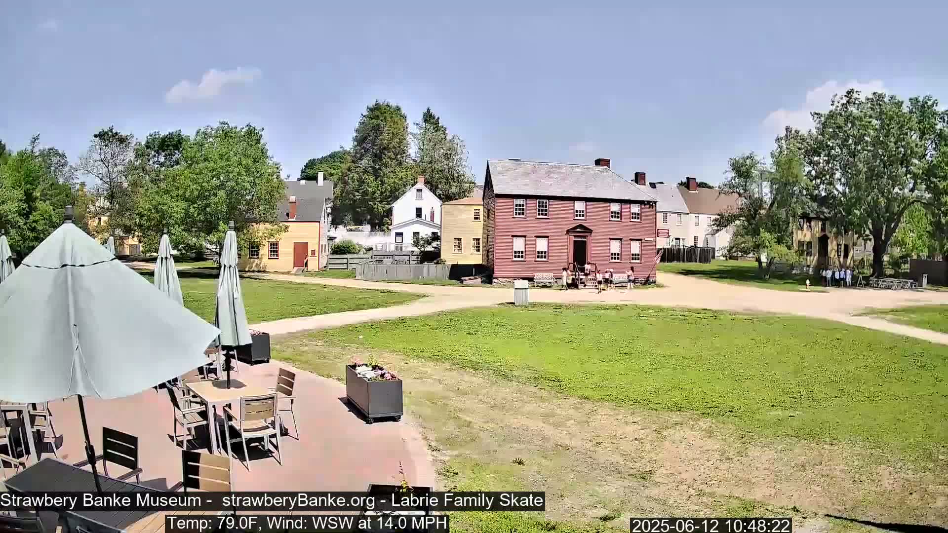 A sunny outdoor scene shows several historic-looking buildings and a grassy area with outdoor seating, under a mostly clear sky.