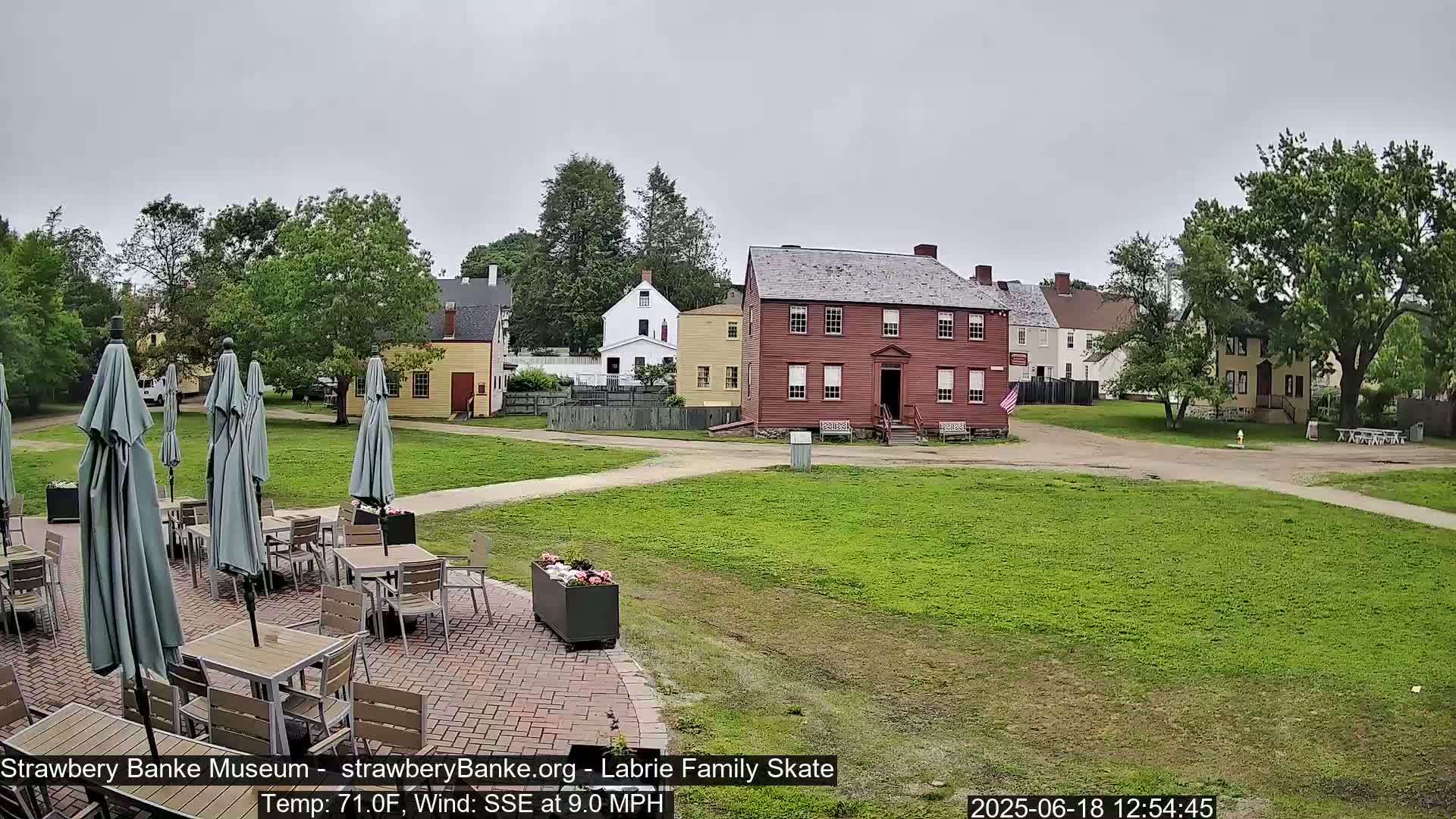A patio with tables and umbrellas overlooks a grassy area and several historic-looking buildings on an overcast day.