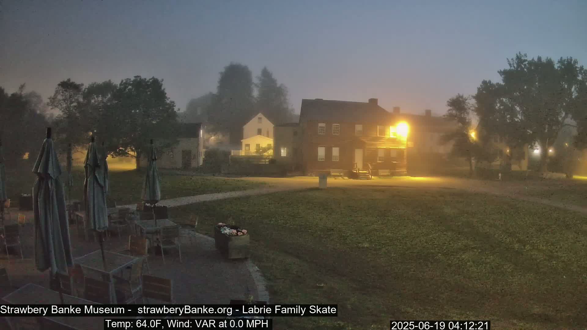 Several closed patio umbrellas surround empty tables and chairs on a grassy lawn near dimly lit, fog-shrouded buildings at night.