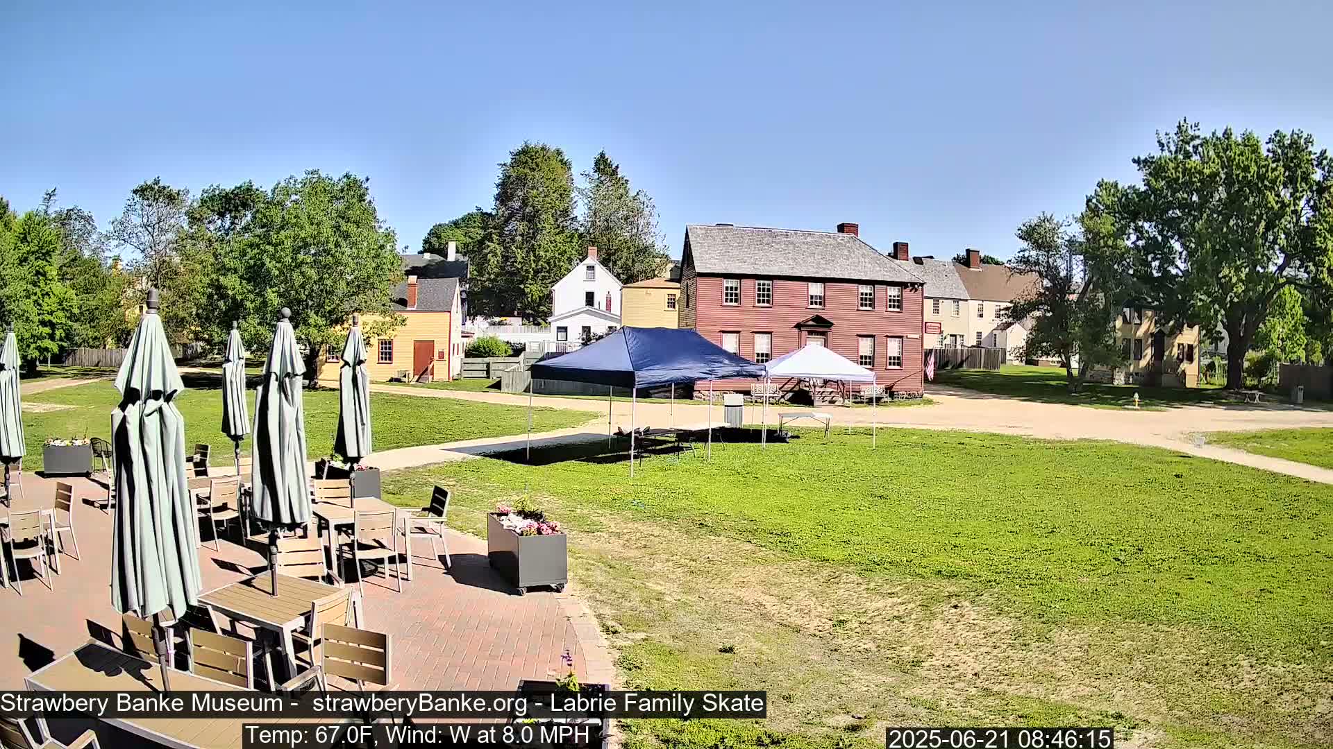 A sunny outdoor scene shows a patio with tables and umbrellas in front of several historic-looking buildings, with a grassy area beyond.