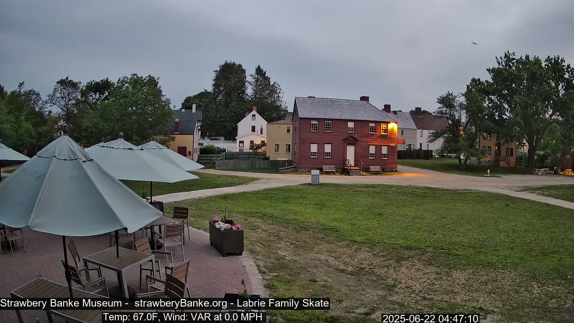 An outdoor patio with several umbrellas and tables overlooks a grassy area and several historic-looking buildings under an overcast sky.