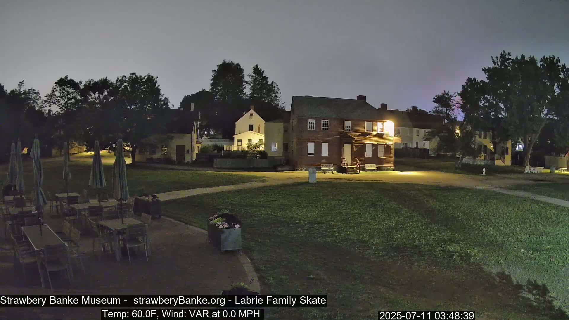 A nighttime view of a grassy area with several buildings, outdoor tables and chairs, and closed umbrellas.