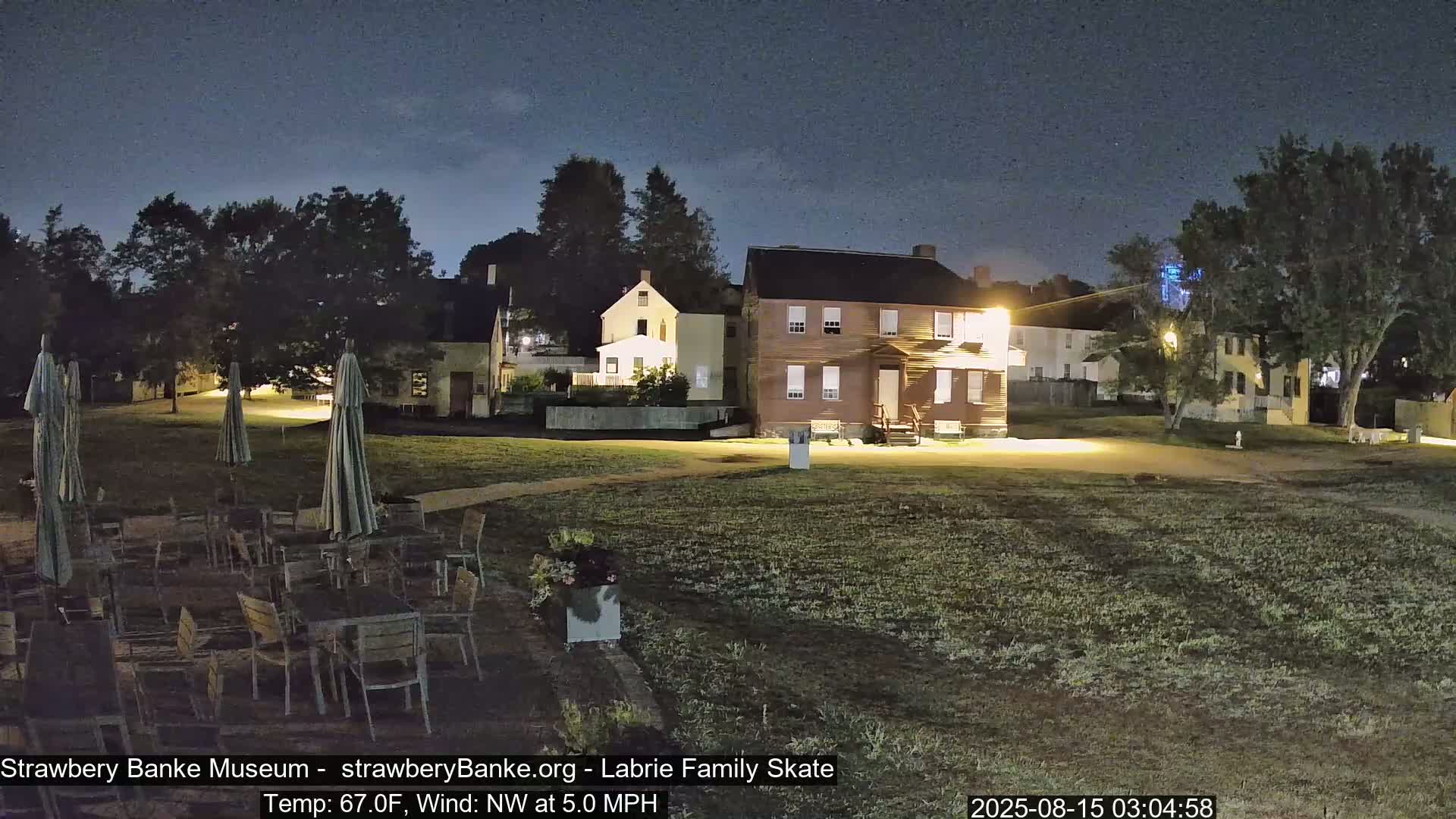 A nighttime outdoor scene shows several illuminated historical buildings surrounded by a grassy area with patio furniture and umbrellas under a partly cloudy sky, with a northwest wind of 5 mph and a temperature of 67°F.
