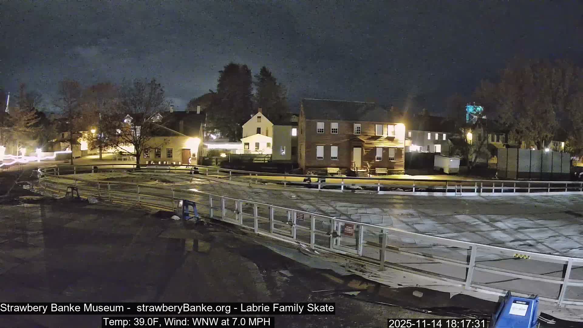 The image shows a clear, dark night over a historical village, with an un-iced rink or prepared area in the foreground enclosed by a white fence, and several warmly lit historic buildings and bare trees in the background.