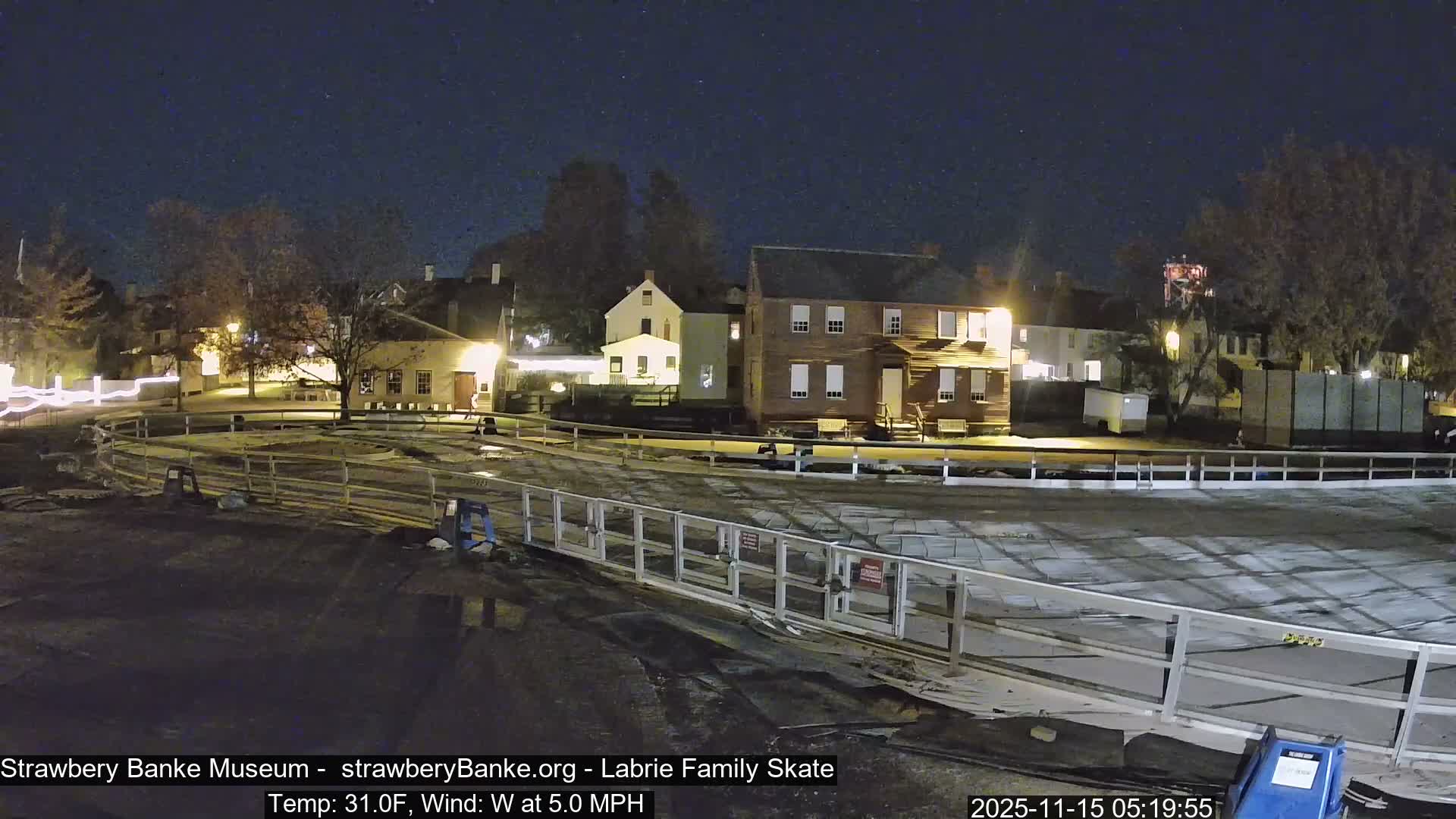 A clear, cold night at 31.0°F with a 5.0 MPH wind illuminates historic colonial-style buildings in the background, overlooking a fenced outdoor area with damp ground, likely an ice rink under preparation.