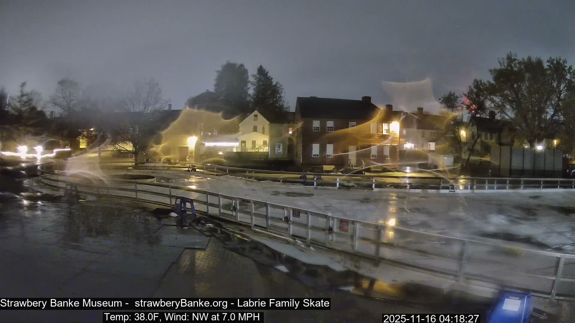 The image captures a wet outdoor scene at night, featuring a damp paved area and a fence in the foreground with bright, streaky light reflections, and illuminated buildings in the background under an overcast sky, suggesting rainy or misty weather.