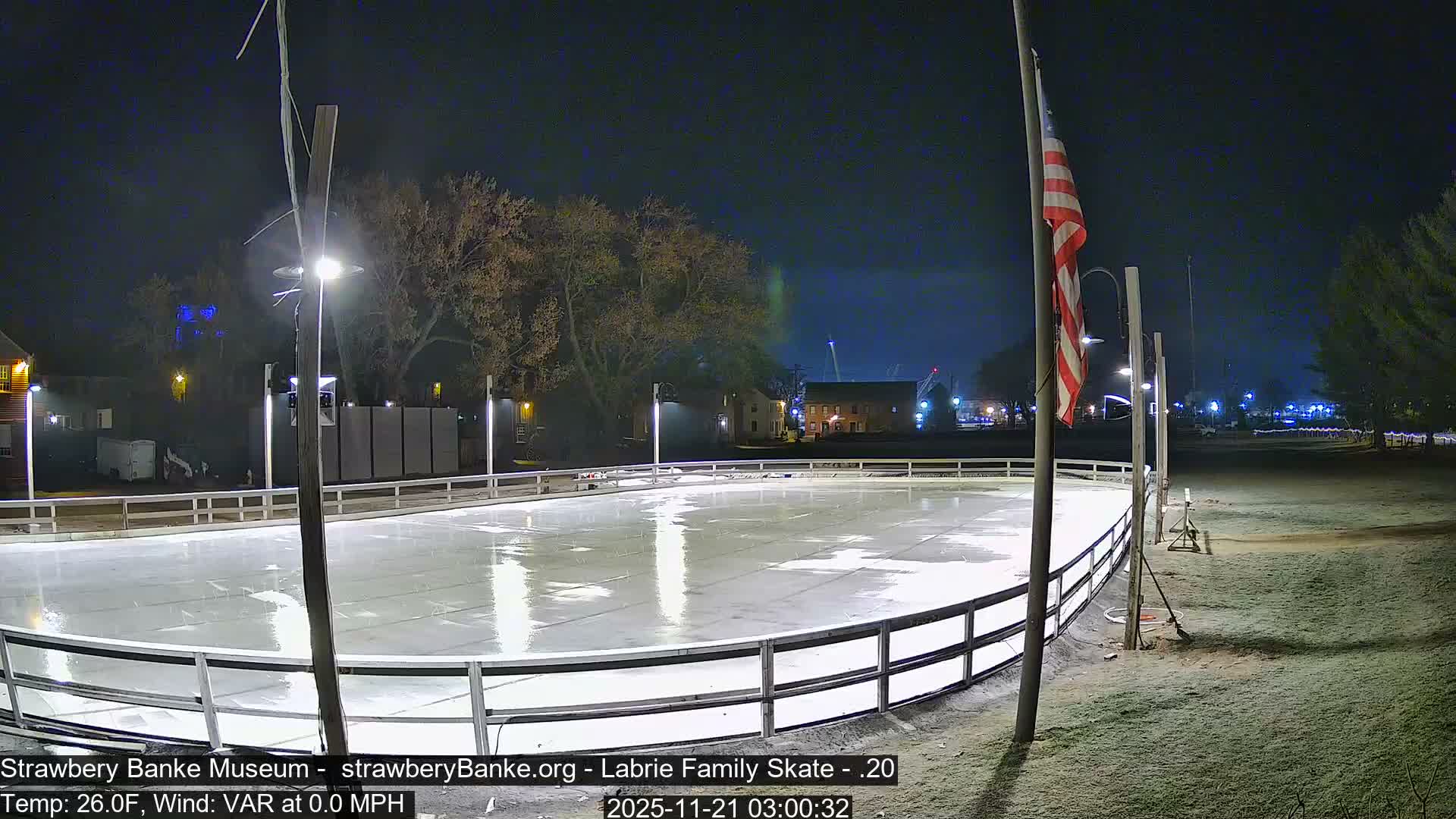 An outdoor ice rink, gleaming under artificial lights and surrounded by a white barrier, lies empty on a clear, cold night with an American flag flying from a pole to the right.