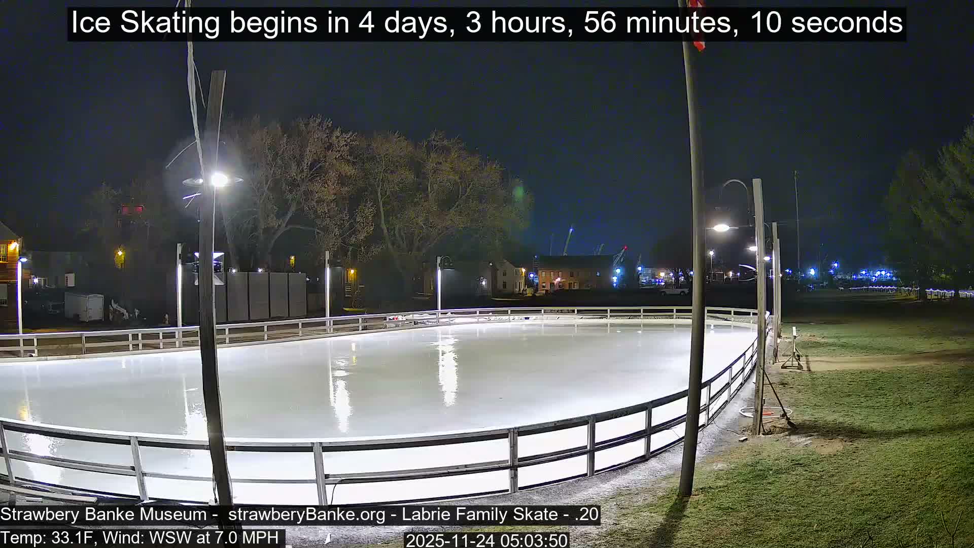 An empty, brightly lit outdoor ice rink, surrounded by a white barrier, gleams under clear night skies with bare trees and distant buildings in the background.