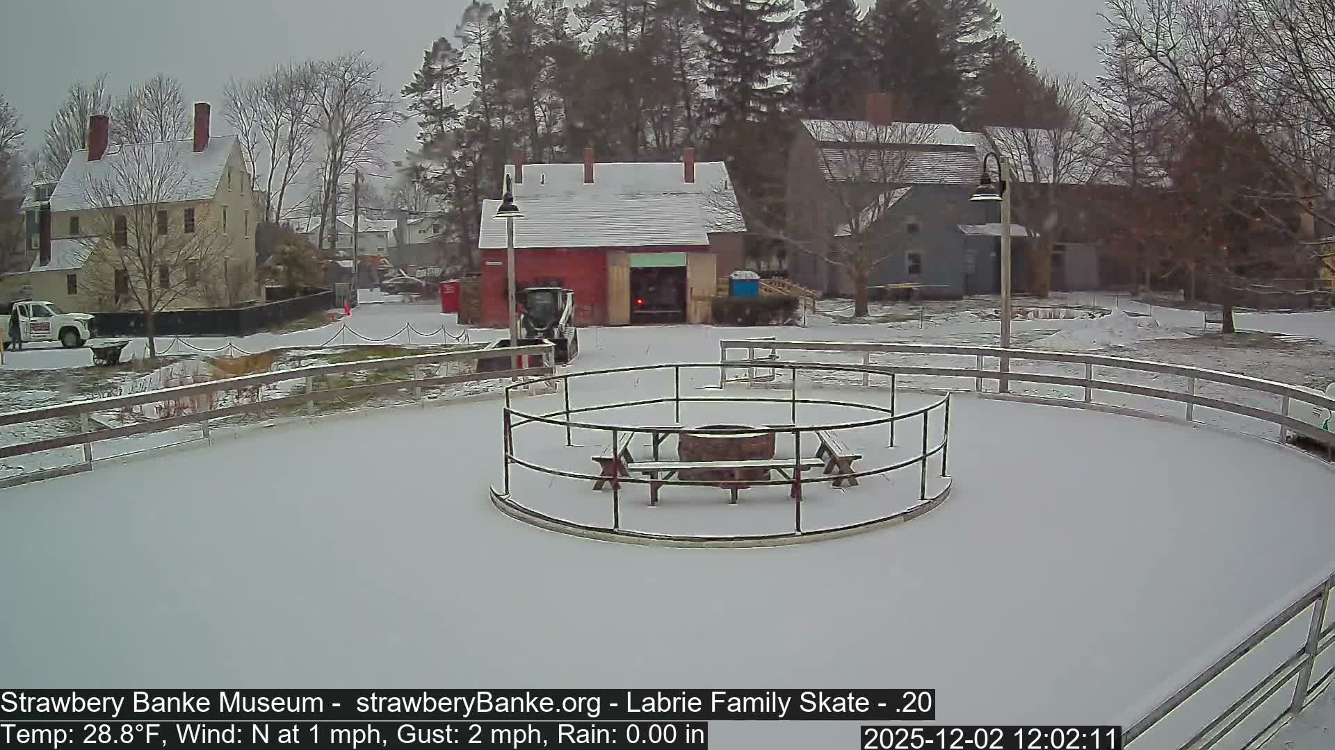 An outdoor historical village scene is covered in a fresh layer of snow under an overcast sky, featuring a circular skating rink or patio with a central fire pit and picnic tables, surrounded by buildings and bare trees.