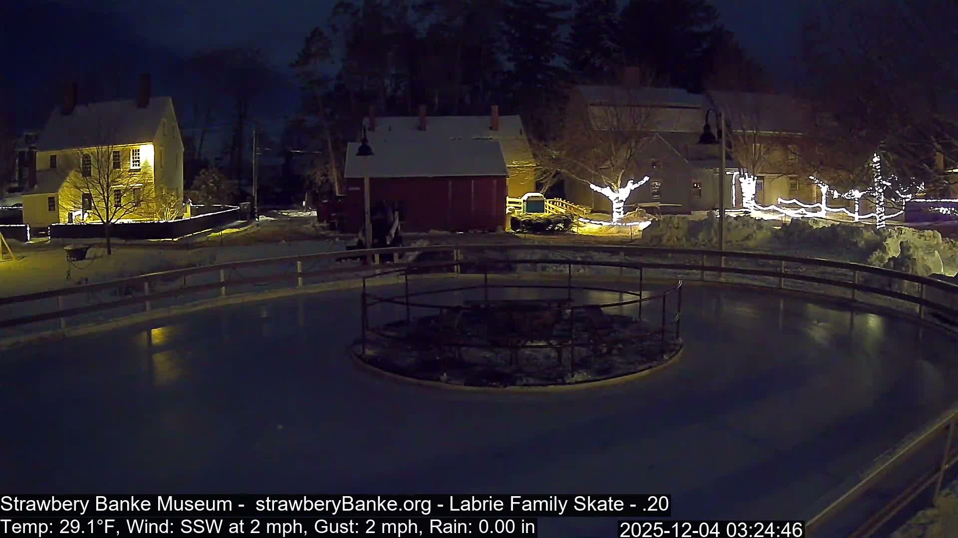 An illuminated outdoor ice skating rink is surrounded by snow-covered historic buildings and trees adorned with white lights on a clear, cold winter night.