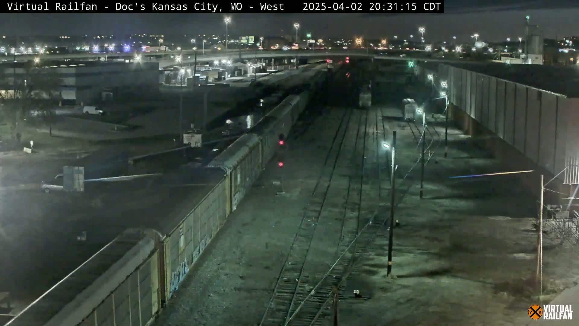 A long freight train is parked at night next to a high wall near a highway, under a clear sky.