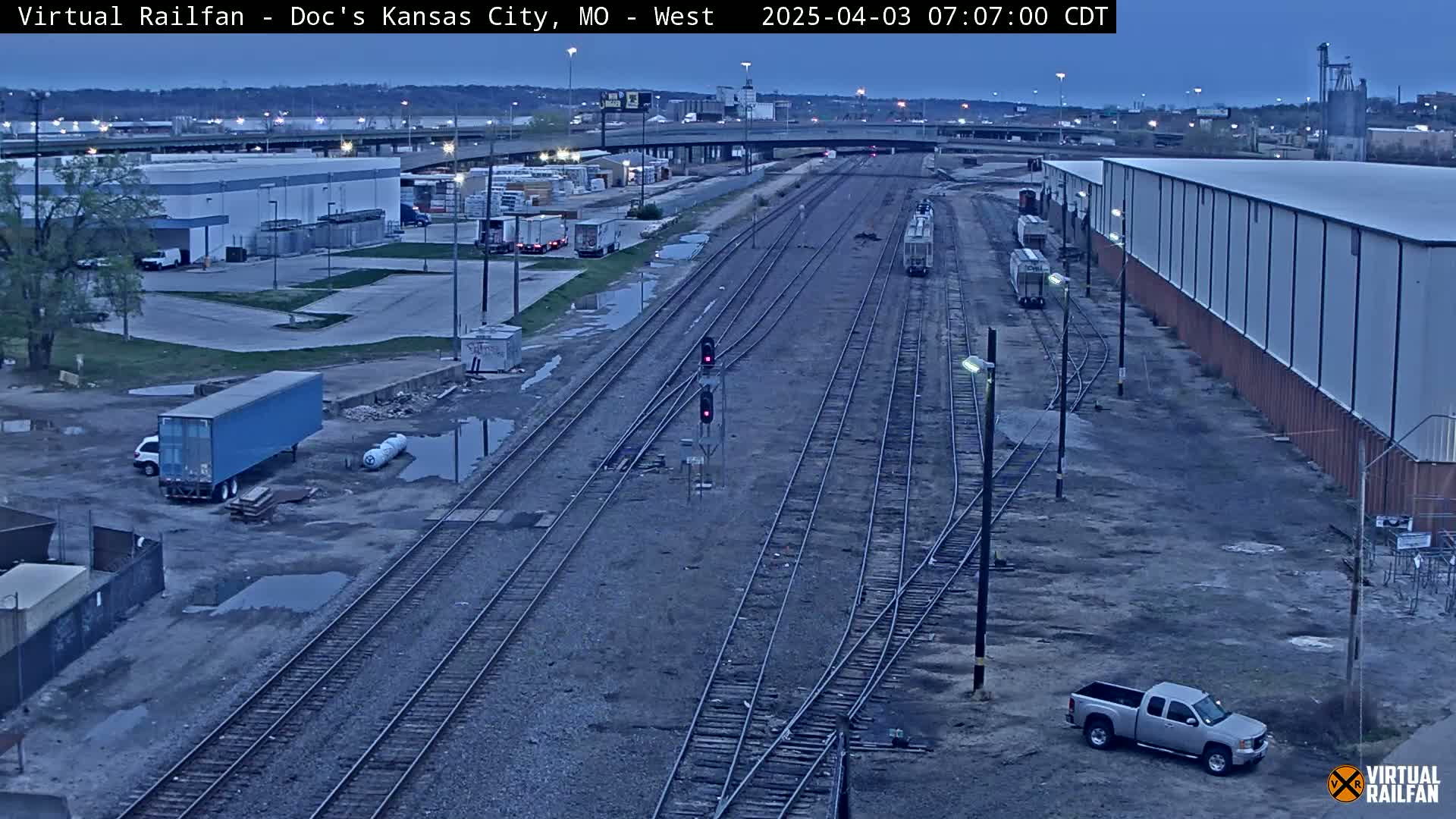 A high-angle, long shot shows a railway yard with several train cars, trucks, and buildings under a twilight sky.