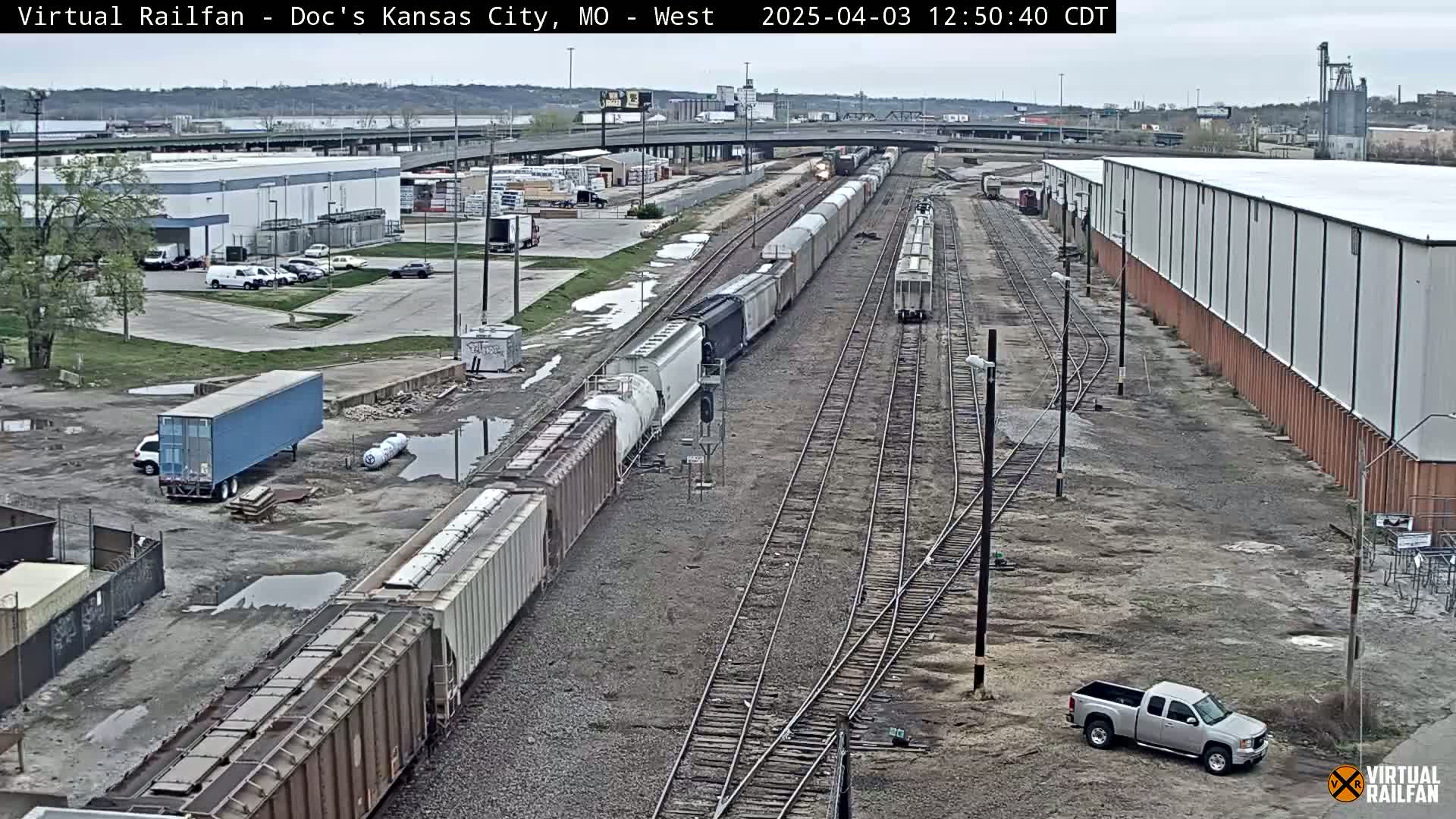 A long freight train is moving along a set of railway tracks in an overcast outdoor setting, with several other vehicles visible in a nearby parking lot and industrial area.