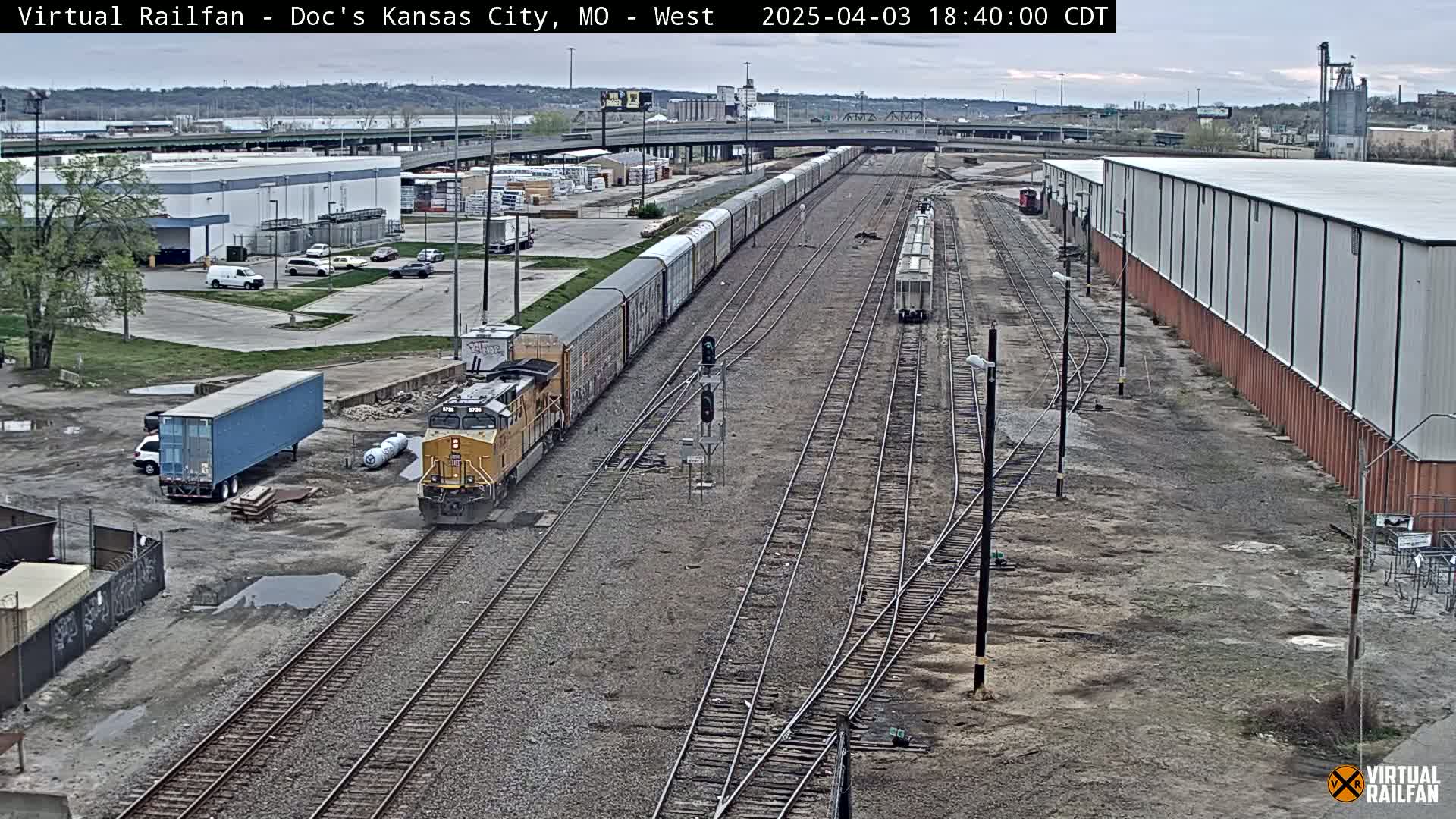 A long freight train and a shorter freight train are visible on a rail yard under an overcast sky.