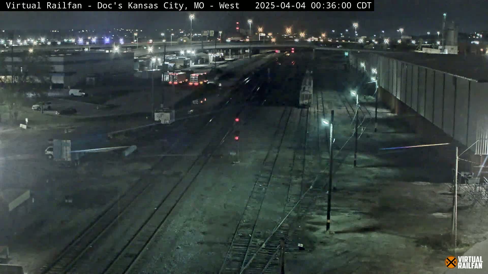 A nighttime aerial view of a train yard with several rail lines, some parked vehicles, and a long building under mostly clear skies.