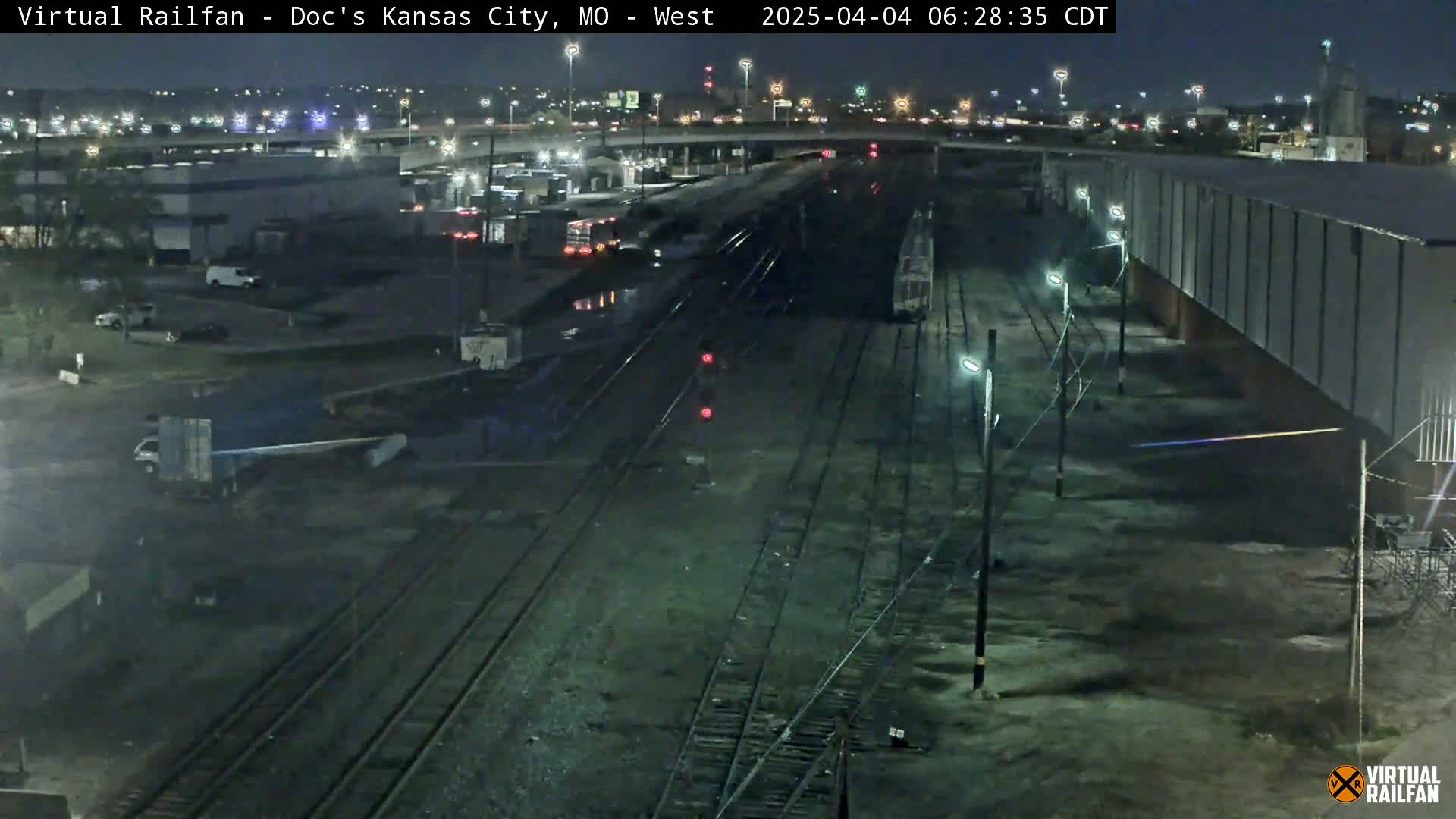 A nighttime, overhead view of railroad tracks and industrial buildings, with a train visible in the distance and some vehicles parked nearby.