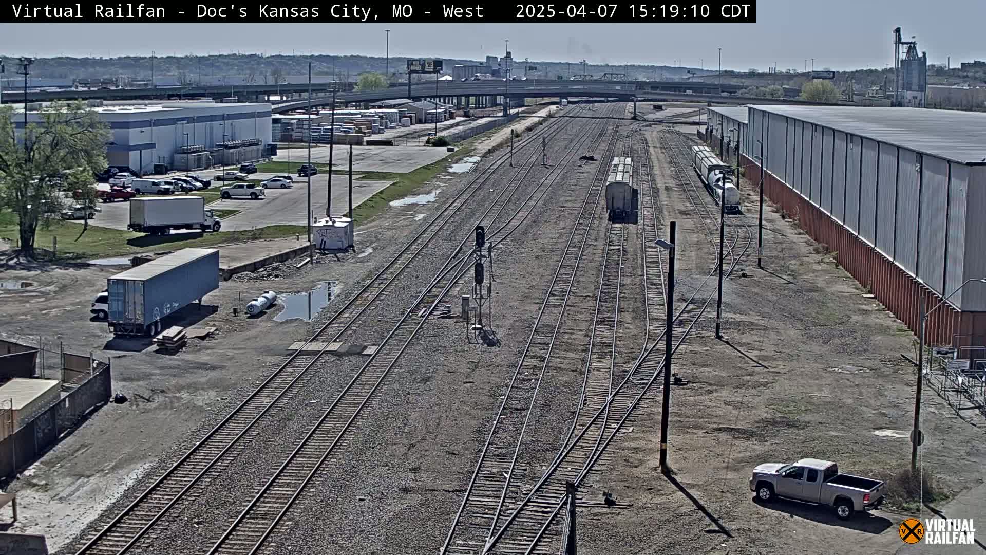 A high-angle view shows a railroad yard with multiple tracks, several parked vehicles, industrial buildings, and a partly cloudy sky.