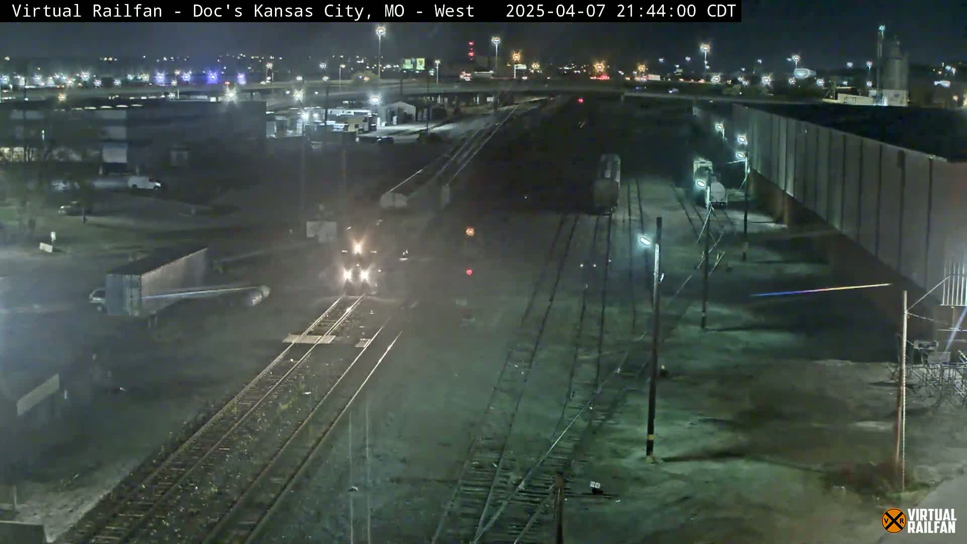 A nighttime view of a train moving along railroad tracks near an industrial area, under clear skies.