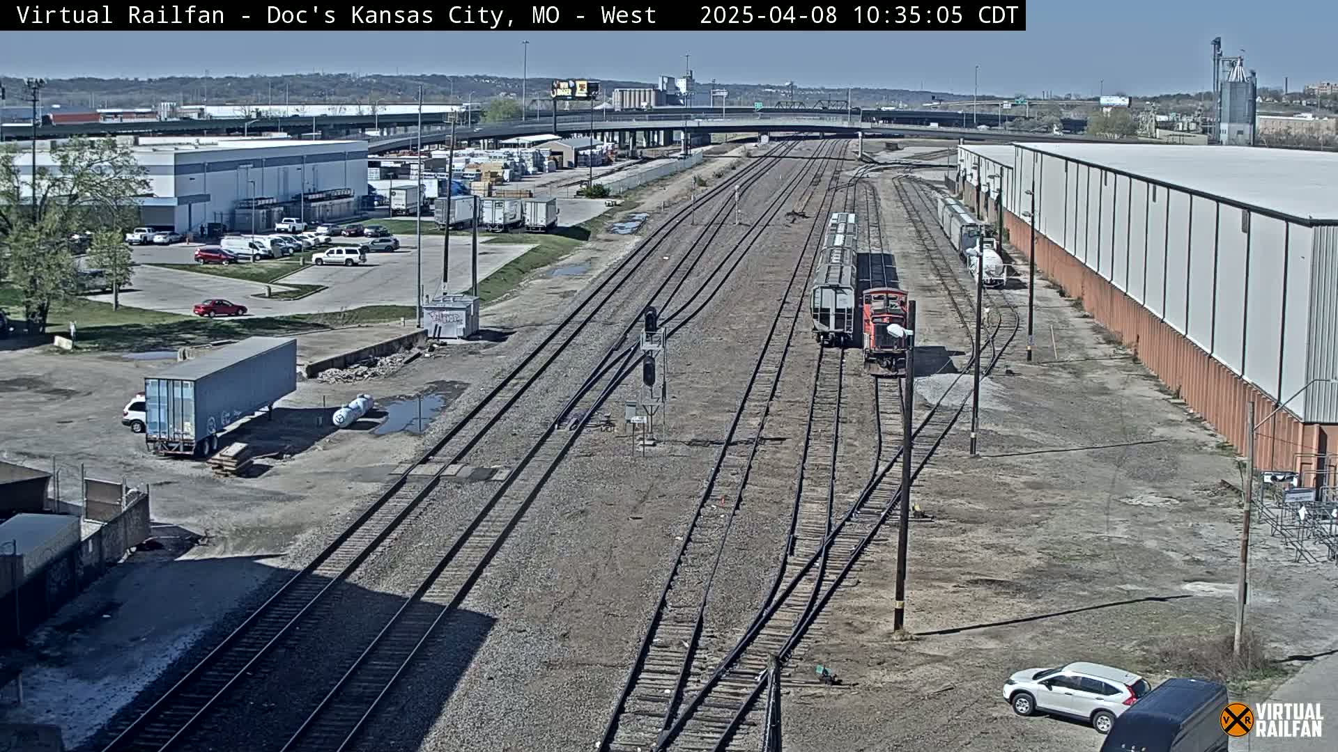 A view of a train yard with several rail lines, industrial buildings, and parked cars under a clear, sunny sky.