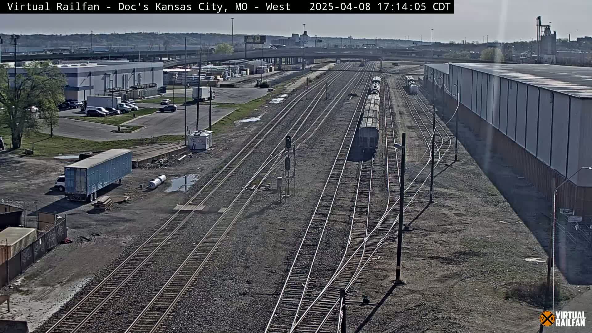 A freight train sits on a railway track next to industrial buildings and parking lots under a partly sunny sky.