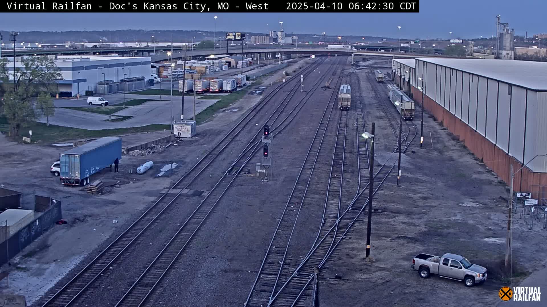 A twilight view of a rail yard with several train cars, trucks, and buildings surrounding a network of train tracks.