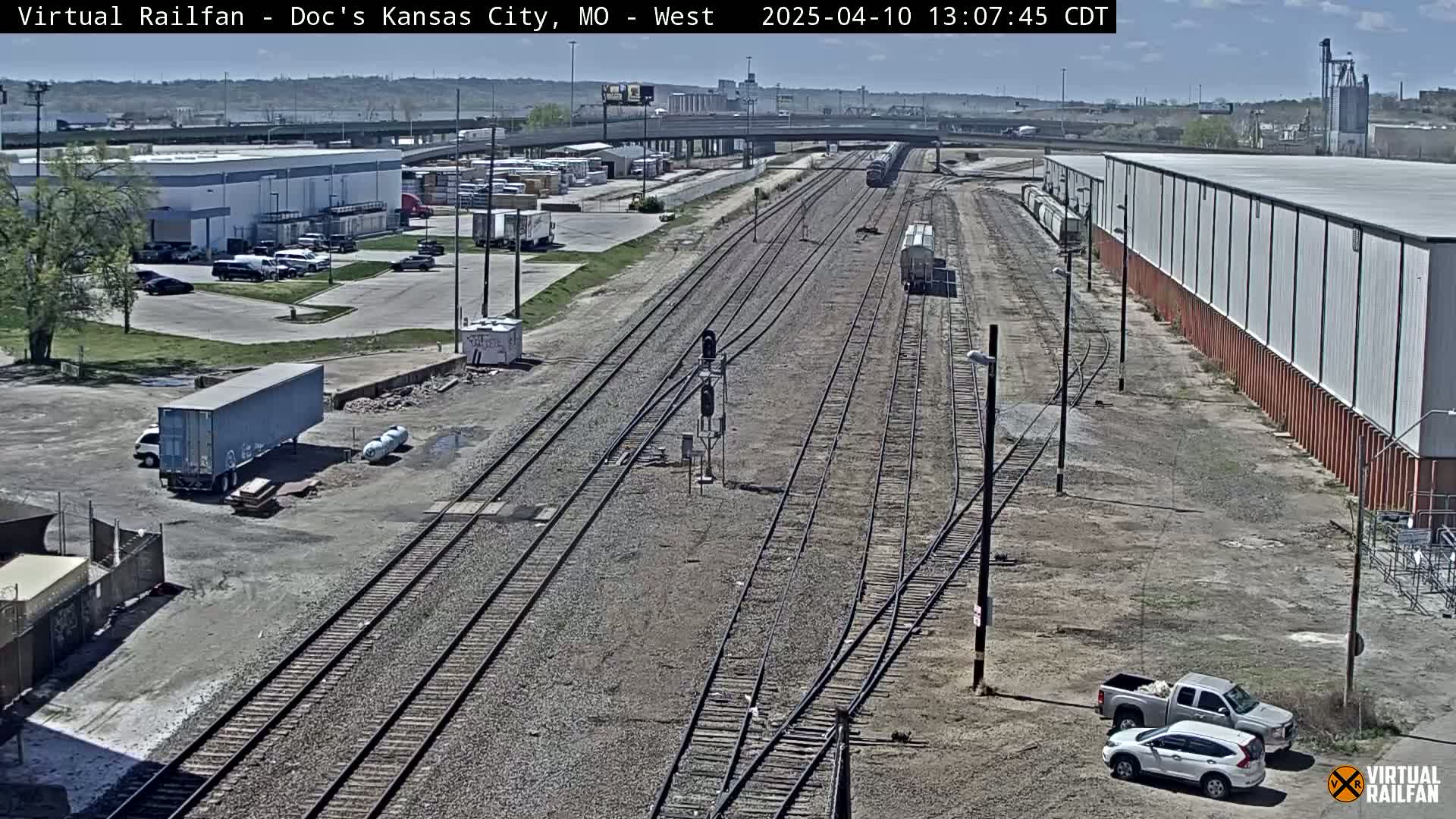 A high-angle view of railroad tracks and industrial buildings on a sunny day, with several vehicles parked nearby.