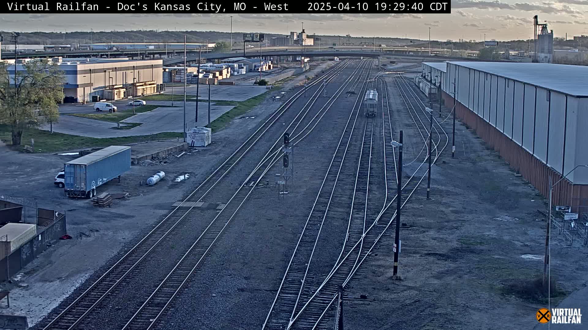 A high-angle, long shot shows a rail yard with several train tracks converging, surrounded by industrial buildings and a few vehicles under a partly cloudy evening sky.