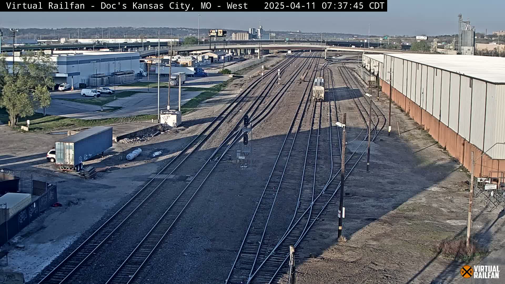 A sunny view of a railway yard with several train tracks converging, surrounded by industrial buildings and a few parked vehicles.
