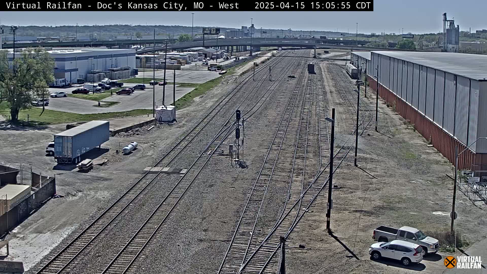 A high-angle view of railroad tracks on a sunny day, with several buildings, vehicles, and a semi-trailer truck visible in the surrounding area.