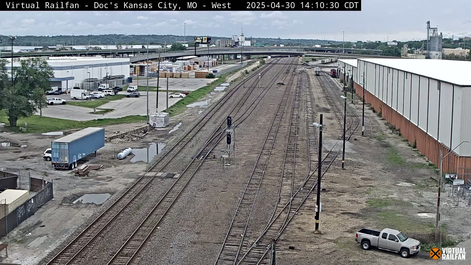 A view of railroad tracks and industrial buildings on an overcast day, with a few vehicles visible in parking lots and along the tracks.