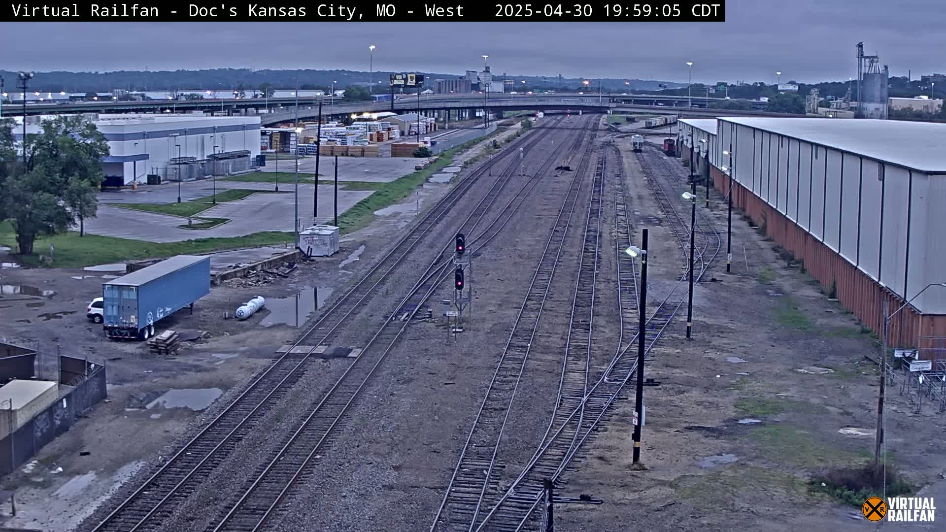 A view of several sets of train tracks next to industrial buildings and a truck, under an overcast sky.