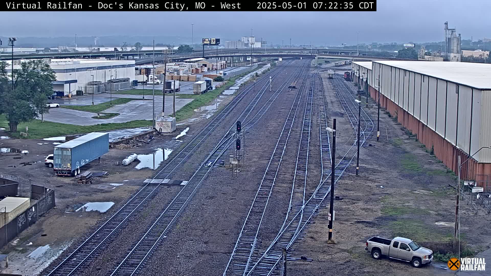 A view of railroad tracks and industrial buildings on an overcast day, with puddles on the ground and a pickup truck parked nearby.