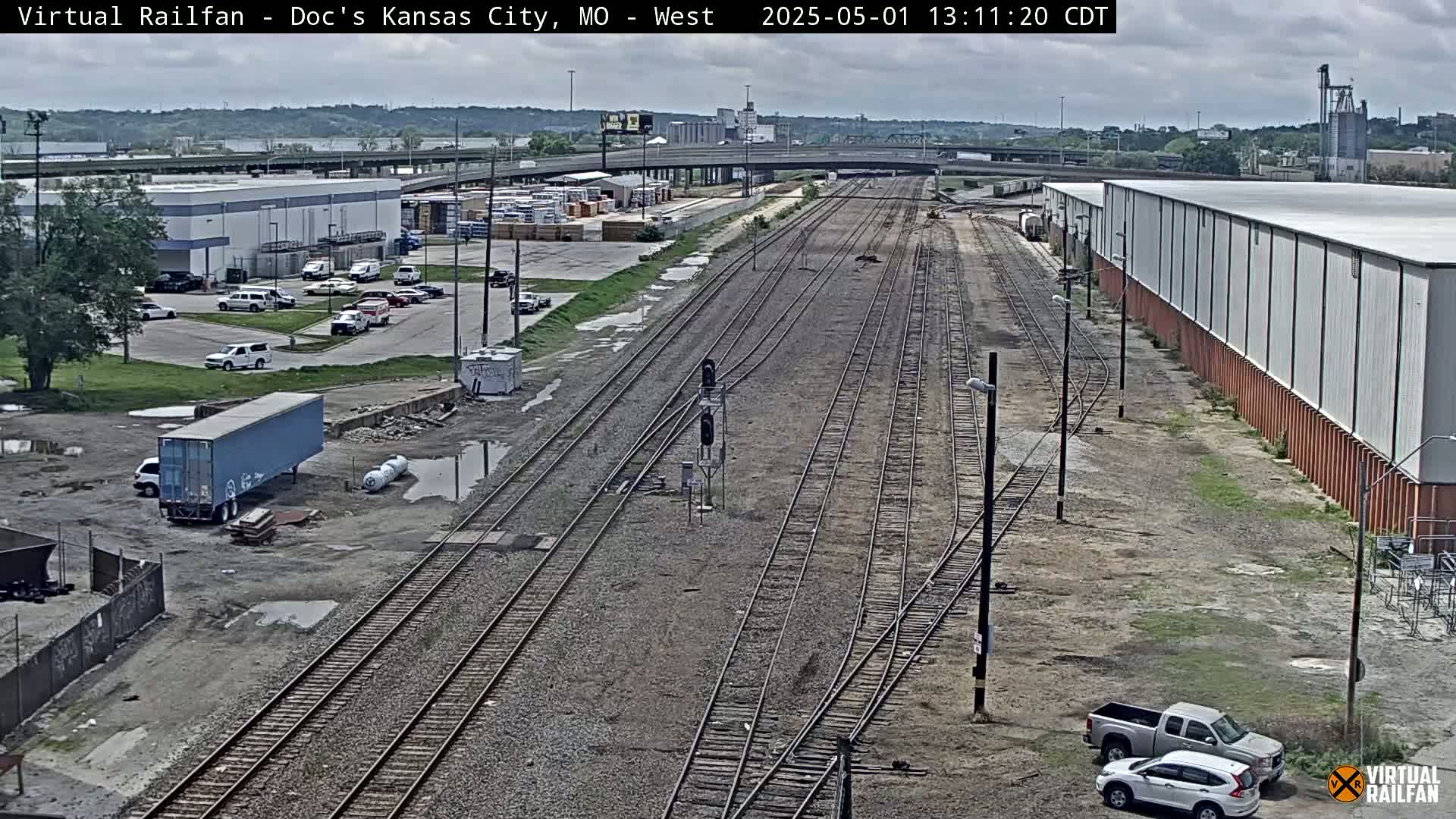 A high-angle, long shot shows several sets of train tracks extending into the distance alongside industrial buildings and a parking lot under a partly cloudy sky.