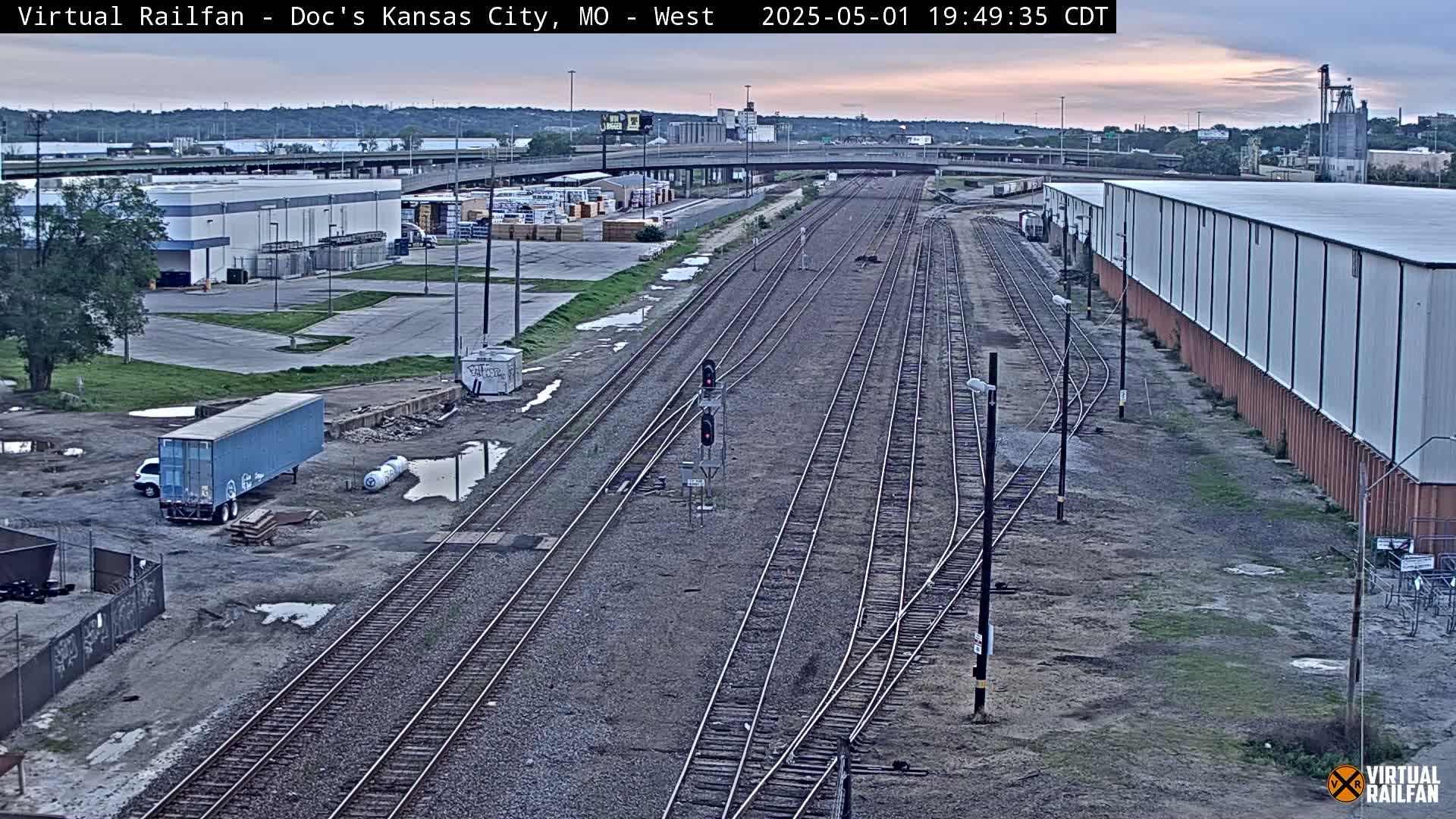 A twilight view of railroad tracks and industrial buildings with puddles of water on the ground.